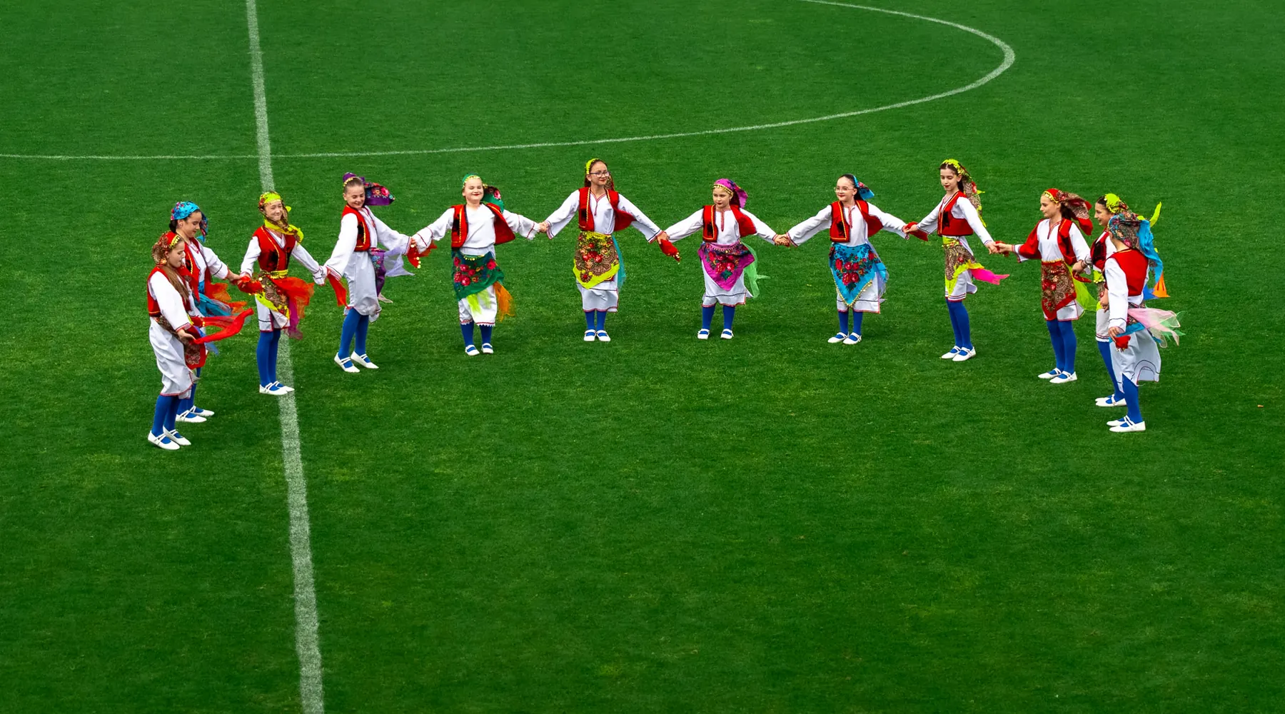 Un círculo de chicas con blusas blancas, chalecos rojos y delantales bordados se dan la mano en un campo de fútbol