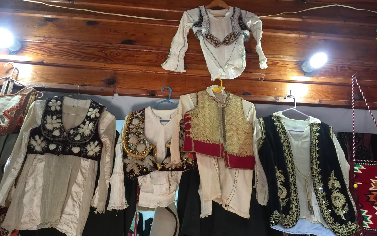 Blusas y chalecos bordados en crema, negro, rojo y dorado cuelgan de perchas bajo un techo de madera