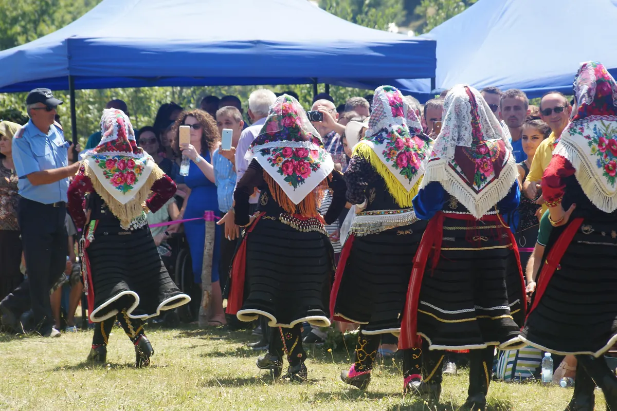 Mujeres con faldas plisadas negras, fajas rojas y pañuelos florales con flecos dorados, vistas de espaldas, bailan sobre la hierba mientras una multitud observa