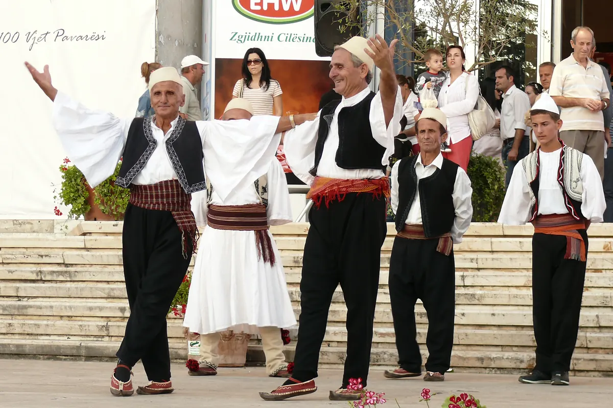 Hombres mayores con camisas blancas, chalecos negros y gorros de fieltro blanco bailan con los brazos extendidos ante una escalinata flanqueada de flores