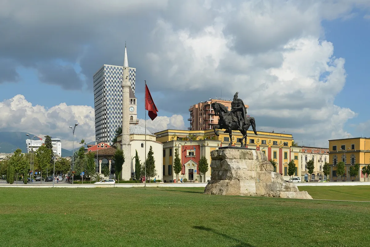 La estatua ecuestre de bronce de Skanderbeg sobre un pedestal de piedra, con el minarete de la Mezquita Et'hem Bey y la torre del reloj alzándose tras los árboles