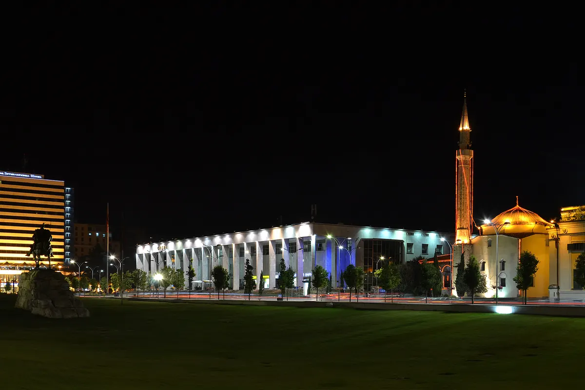 La plaza Skanderbeg de noche, con un edificio blanco de columnas iluminado en el centro, una torre de hotel iluminada a la izquierda y la cúpula dorada y el minarete de la mezquita Et'hem Bey a la derecha