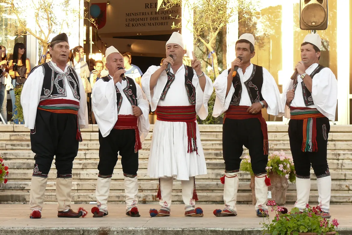 Cinco hombres con camisas blancas, chalecos bordados y fajas a rayas cantan ante micrófonos en un escenario al aire libre